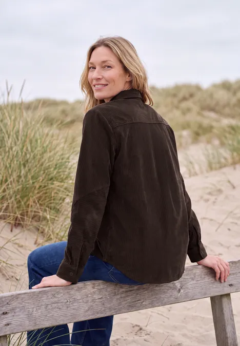 Langarm Hemdbluse aus Cord in Unifarbe truffle brown Frau mit blonden Haaren in brauner Jacke sitzt auf einer Bank vor Dünen, lächelt zur Kamera.