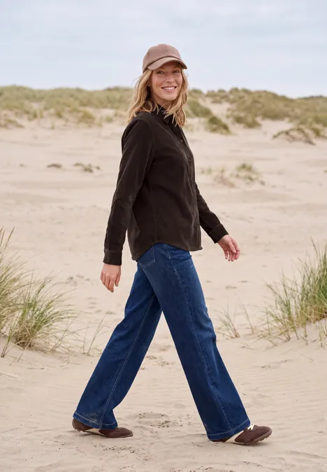 Langarm Hemdbluse aus Cord in Unifarbe truffle brown Frau in schwarzer Jacke, Jeans und Mütze spaziert lächelnd über Sanddünen am Strand.