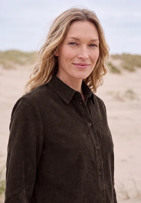 Langarm Hemdbluse aus Cord in Unifarbe truffle brown Eine Frau mit hellem Haar und dunklem Hemd steht lächelnd am Strand vor Dünen.
