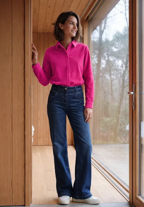 Langarm Hemdbluse aus Cord in Unifarbe pink flash Frau in pinker Bluse und blauen Schlagjeans steht lächelnd in einem hellen Holzraum am Fenster.