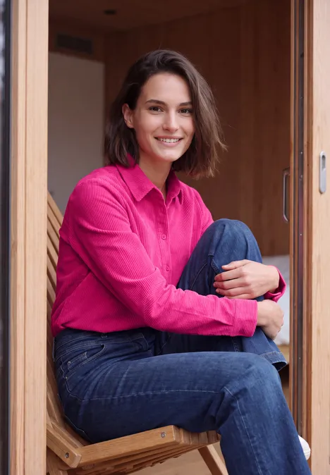 Langarm Hemdbluse aus Cord in Unifarbe pink flash Eine Frau mit pinkem Hemd und Jeans sitzt im Holzrahmen und lächelt.