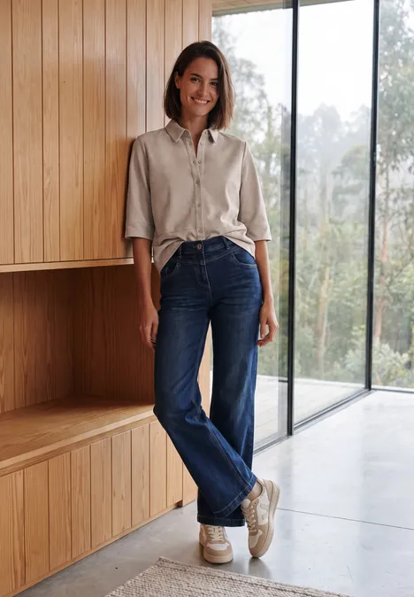 Kurzarm Hemdbluse in Unifarbe cashmere beige Frau in beigem Hemd, Jeans und Sneakers steht in hellem, modernem Raum neben einer großen Fensterfront.