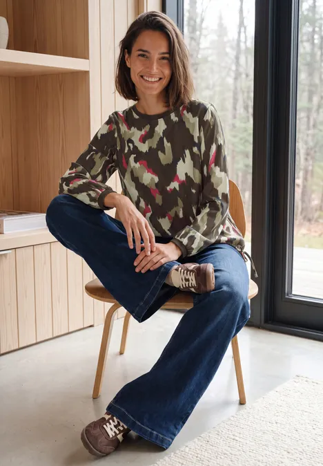 Blouse met lange mouwen, ronde hals en print pink flash Een vrouw zit glimlachend op een stoel bij een groot raam, draagt een camouflage trui en een spijkerbroek.