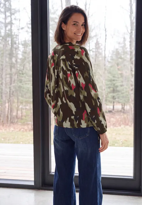Blouse met lange mouwen, ronde hals en print pink flash Een lachende vrouw in een camouflage trui en spijkerbroek staat voor een groot raam en kijkt over haar schouder heen.