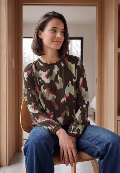 Blouse met lange mouwen, ronde hals en print pink flash Vrouw zit in moderne houten ruimte, draagt groene camouflage-trui met roze accenten en blauwe jeans, glimlachend opzij kijkend.
