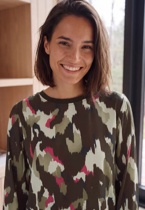 Blouse met lange mouwen, ronde hals en print pink flash Jonge vrouw met kort donker haar, glimlacht in een camouflagetrui bij een helder raam.