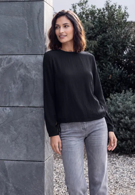 Langarm Blusenshirt mit Smokdetails urban dark blue Eine Frau in schwarzer Bluse und grauer Jeans lehnt im Freien an einer Steinwand.