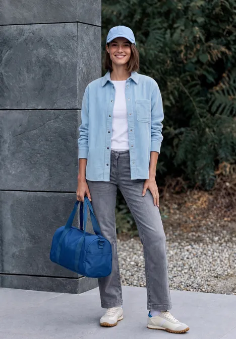 Frau in Jeanshemd, weißem T-Shirt, grauen Hosen und Sneakern mit blauer Reisetasche im Freien.