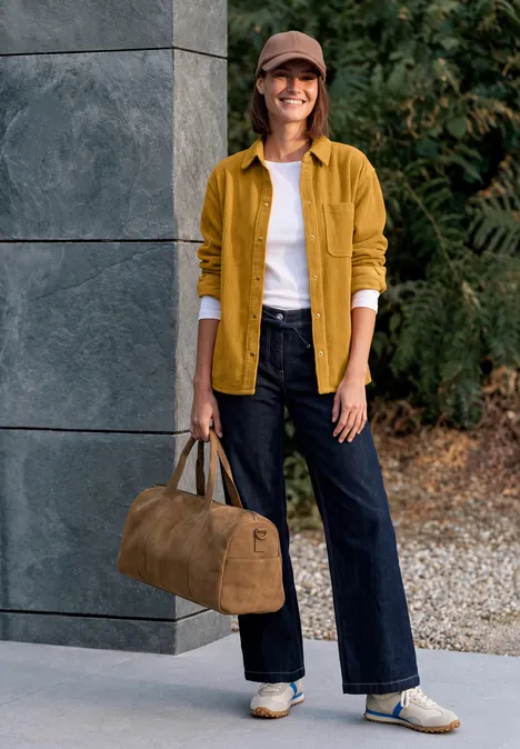 Frau in gelber Jacke, weißem Shirt, weiter Jeans und Sneakern, hält eine braune Reisetasche und lächelt.