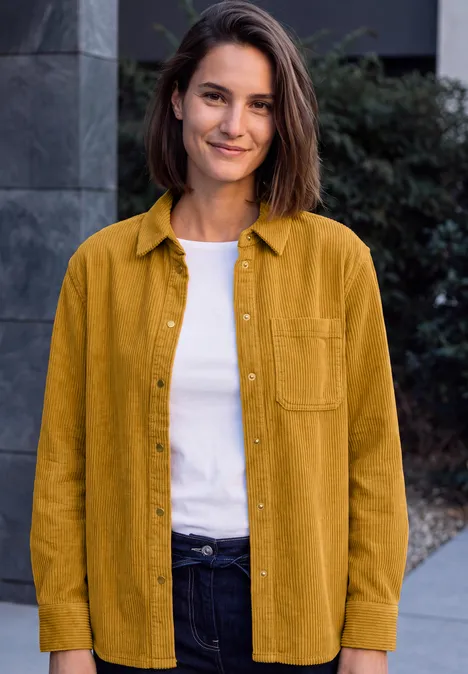 Eine lächelnde Frau vor einem Gebäude trägt ein senfgelbes Hemd, weißes T-Shirt und dunkle Jeans.