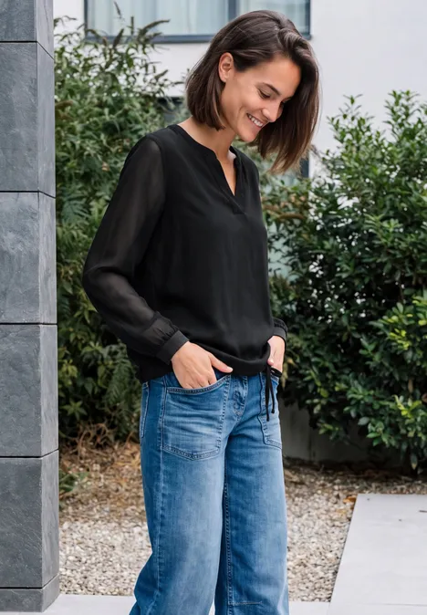 Langarm Bluse mit V-Neck und Strukturmix Black Eine Frau in schwarzer Bluse und blauen Jeans steht lächelnd draußen vor einem Gebäude mit Grün.