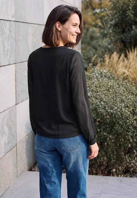 Langarm Bluse mit V-Neck und Strukturmix Black Frau in schwarzer Bluse und Jeans, steht lächelnd seitlich an einer Mauer, im Hintergrund Pflanzen.