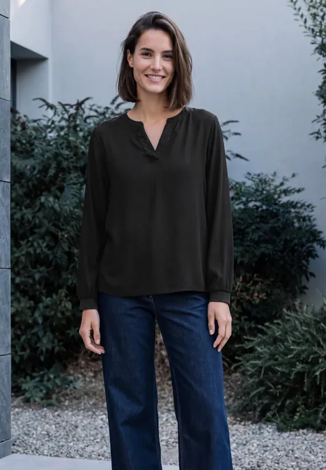 Langarm Tunika mit Split Neck und Detail Black Eine Frau in schwarzer Bluse und Blue Jeans steht lächelnd im Freien vor einer modernen Hausfassade und Büschen.