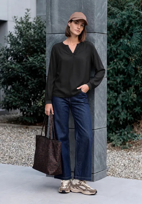 Langarm Tunika mit Split Neck und Detail Black Frau mit Cap, schwarzer Bluse, Jeans und Sneakers posiert mit Tasche vor moderner Hausfassade.