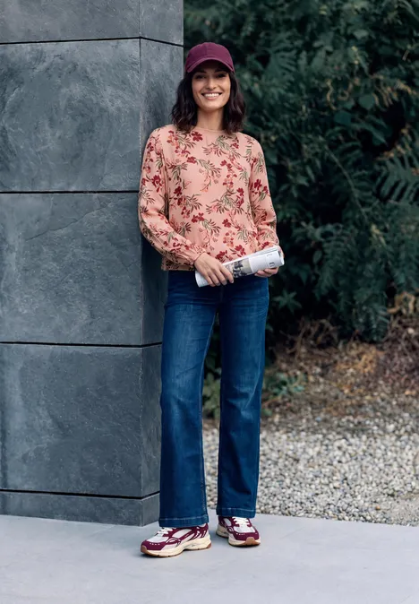 Crepe Bluse mit floralem Muster soft blossom rose Eine Frau mit roter Cap, geblümter Bluse, Jeans und Sneakern lächelt und hält eine Zeitung.