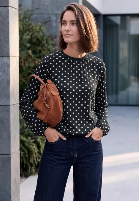 Frau in schwarzem Pullover mit weißen Punkten, Jeans und brauner Tasche steht vor modernem Gebäude.