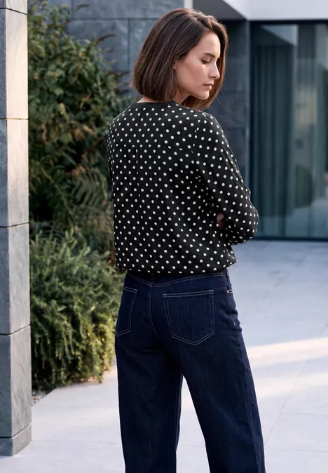 Eine Frau in gepunkteter Bluse und Jeans steht vor modernem Gebäude und blickt zur Seite.