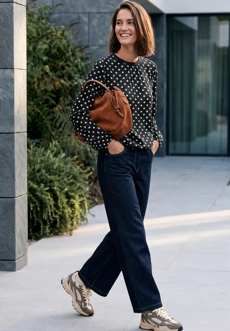 Lächelnde Frau in gepunkteter Bluse, Jeans und Sneakers, die eine braune Tasche an der Schulter trägt und im Freien spaziert.