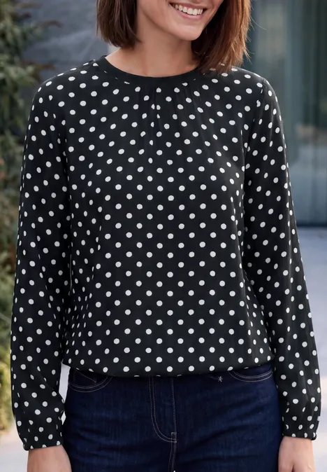 Frau mit schwarzer, langärmeliger Bluse mit weißen Punkten und dunkelblauer Jeans, lächelnd.