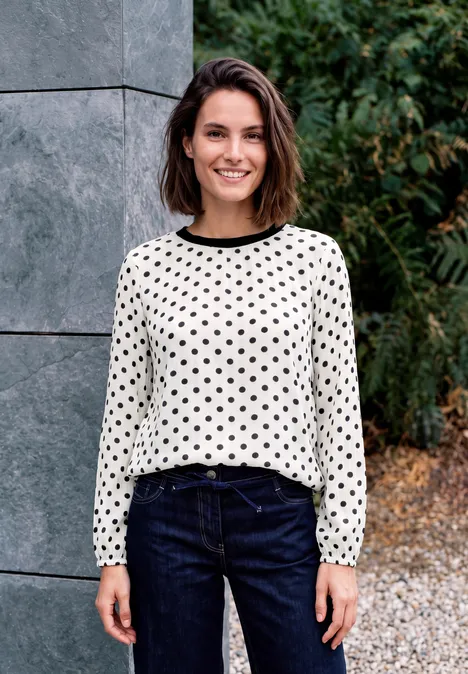 Lächelnde Frau in weißer, schwarz gepunkteter Bluse und blauen Jeans steht draußen vor Natur und Steinwand.