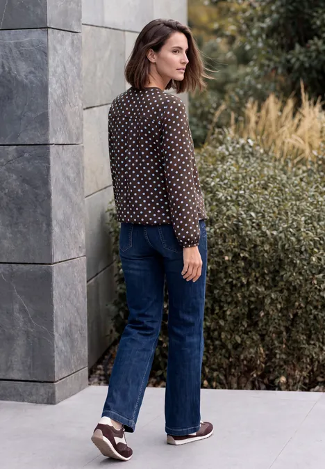 Frau in gepunkteter Bluse und Jeans steht seitlich vor moderner Fassade und Büschen.