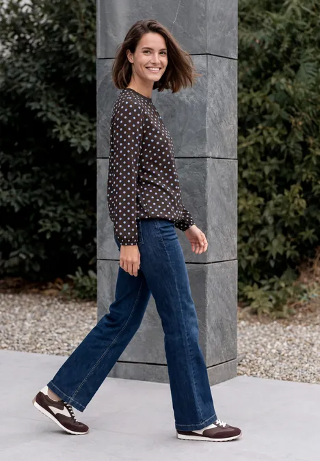 Frau in gepunktetem Pullover und Jeans geht lächelnd an einer grauen Säule vorbei im Garten.