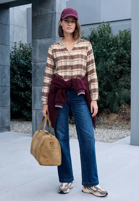 Frau mit lila Cap, kariertem Hemd, Jeans und Beuteltasche vor moderner Gebäudefassade und Pflanzen.