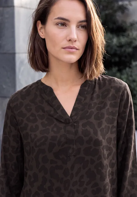 Jonge vrouw met kort bruin haar in een donkerblouse met panterprint, staand tegen een stedelijke achtergrond.