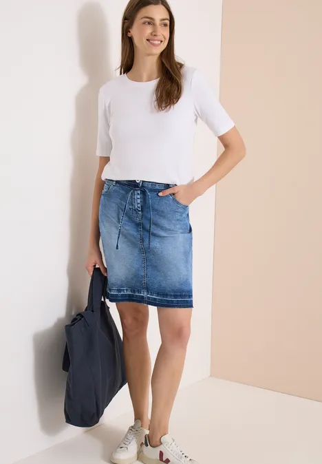 Vrouw voor lichte muur, draagt wit T-shirt, spijkerrok, witte sneakers en houdt een blauwe tas.