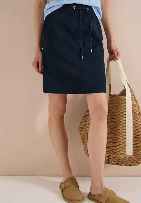 Donkerblauwe rok tot op de knie, lichtblauw shirt, beige slippers en gevlochten rieten tas.