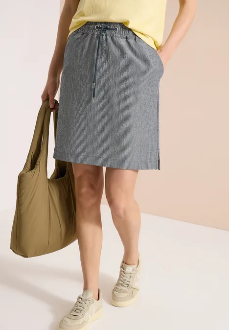 Frau trägt gelbes Top, gestreiften Rock, weiße Sneaker und hält eine khakifarbene Tasche.