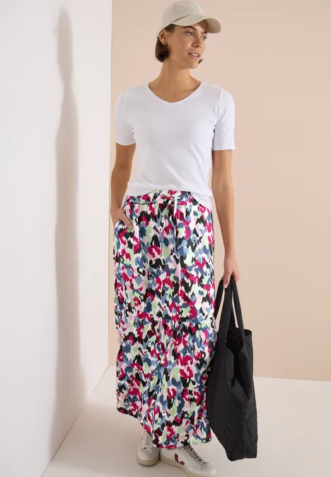 Femme en T-shirt blanc, jupe longue multicolore, casquette blanche, baskets, tenue décontractée avec sac noir.