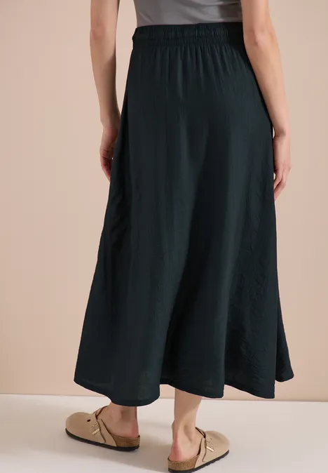 Vrouw van achteren in een zwarte, losvallende midirok en beige sandalen tegen een lichte achtergrond.