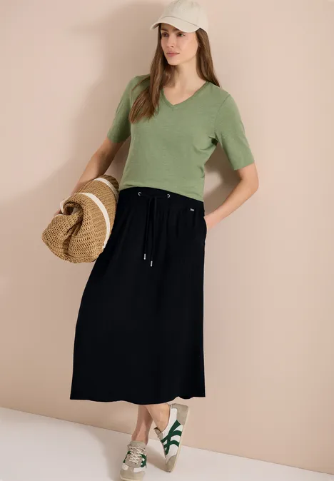 Frau trägt grünes T-Shirt, schwarzen Midirock, Strohtasche und weiße Sandalen mit grünen Details.