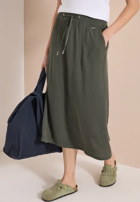 Frau trägt olivgrünen Midirock mit Tunnelzug, weißem Top, grünen Sandalen und blauer Tasche.
