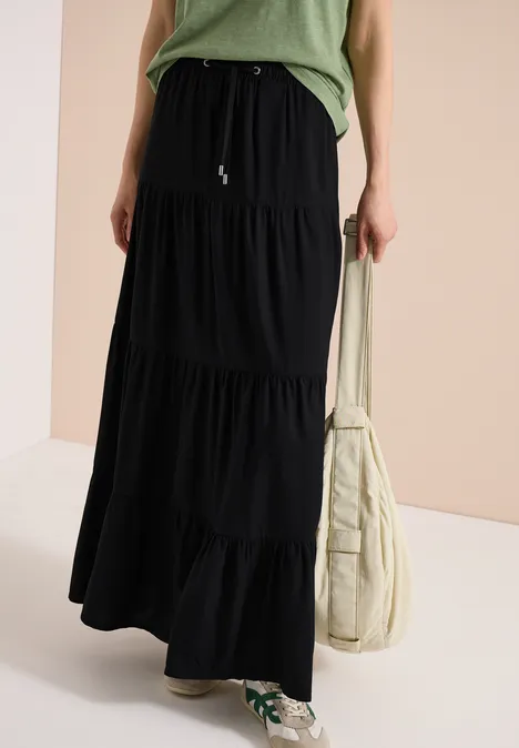 Eine Person trägt einen schwarzen, mehrlagigen Maxirock, grünes Oberteil, helle Schultertasche und grün-weiße Sneaker.