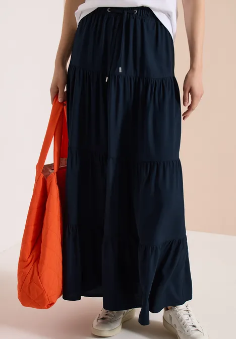 Frau trägt lange, dunkelblaue, gestufte Maxirock, weiße Sneaker und hält eine orangefarbene Tasche.