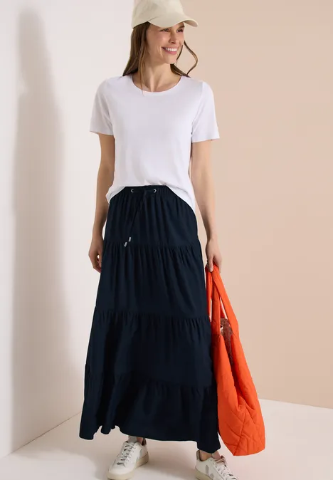 Frau in weißem T-Shirt, schwarzem Stufenrock und grauen Sneakern, hält orange Steppjacke in der Hand.