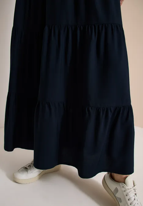 Untere Körperhälfte mit schwarzem, gestuftem Rockteil eines langen Kleides und weißen Sneakern.