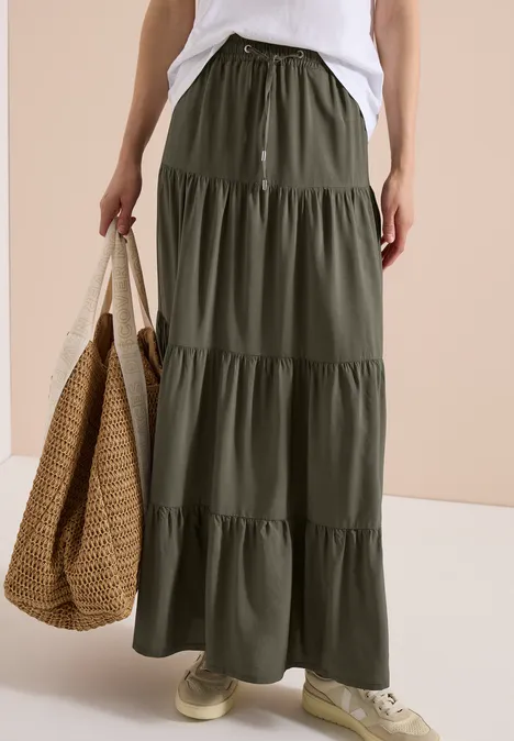 Dame trägt lange, fließende dunkelgrüne Stufenrock, weiße Sneakers und trägt eine große Korbtasche.