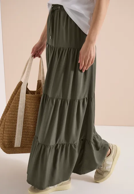 Frau trägt olivgrünen Stufenrock bis zum Knöchel, Basttasche und bequeme Espadrilles-Sneaker.