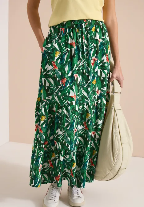 Vrouw in geel top draagt een lange groene rok met bladprint, witte sneakers en beige tas.