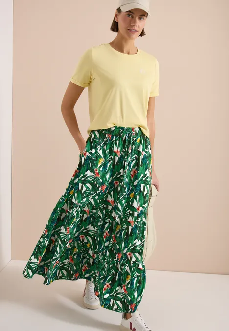 Vrouw in geel T-shirt en lange groene bloemenrok met witte sneakers tegen neutrale achtergrond.