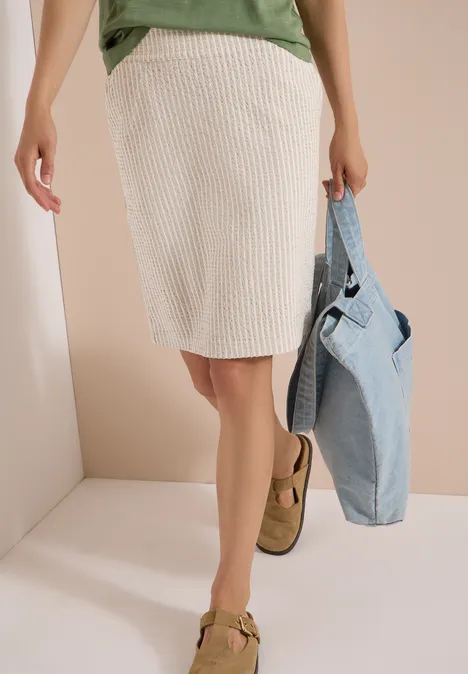 Knielanger Jogg Skirt in Seersucker-Qualität grain beige Frau trägt cremefarbenen, gerippten Rock, grünes Oberteil, hält Jeansjacke und trägt braune Loafer.