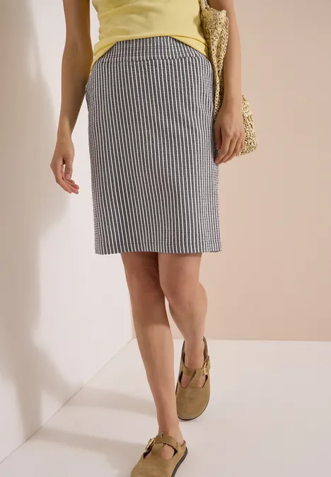Knielanger Jogg Skirt in Seersucker-Qualität brown cacao Frau in knielangem, grau-weiß gestreiftem Rock, gelbem Top und olivgrünen Sandalen vor neutralem Hintergrund.