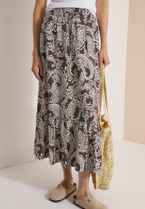 Jupe longue à motifs floraux brown cacao Jupe midi marron à motif cachemire avec volants, portée avec mules beiges et sac en paille.