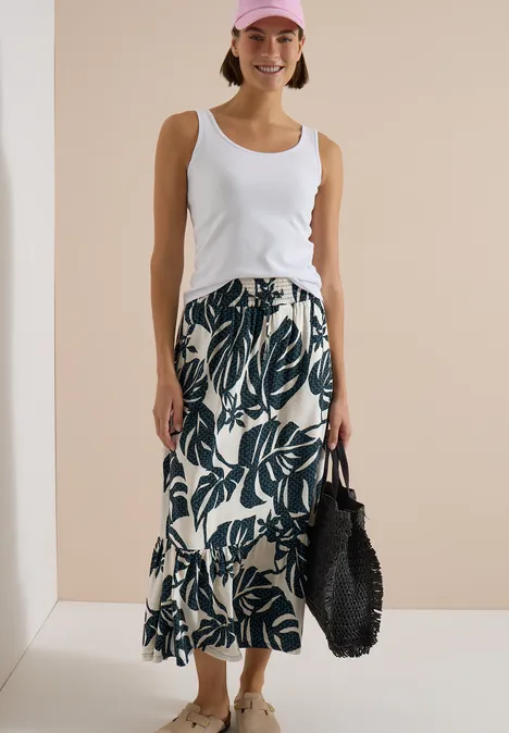 Femme en débardeur blanc et jupe crème à motifs de feuilles vertes, tenant un sac noir.