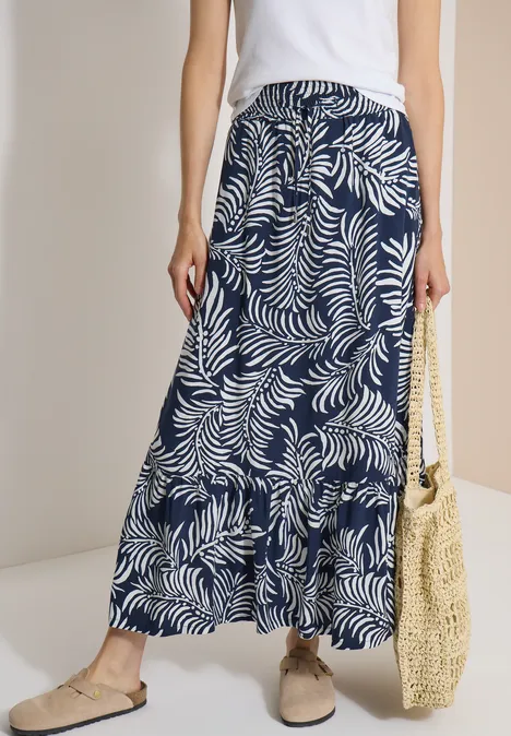 Maxi-Rock mit Blättermuster universal blue Dame trägt weiße Bluse, lange dunkelblaue Rock mit weißem Blattmuster, Basttasche und bequeme Schuhe.