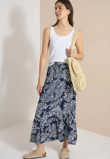 Maxi-Rock mit Blättermuster universal blue Frau in weißem Top und blau-weiß gemusterter Maxirock trägt Basttasche und Sandalen vor zweifarbigem Hintergrund.