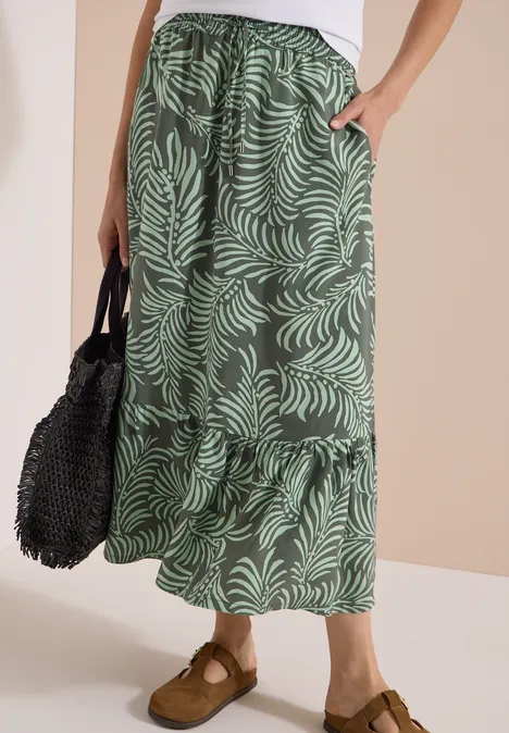 Maxirok met bladmotief khaki Vrouwelijk onderlijf met groene, wijdvallende maxi-rok met bladprint, zwarte tas en bruine mules.